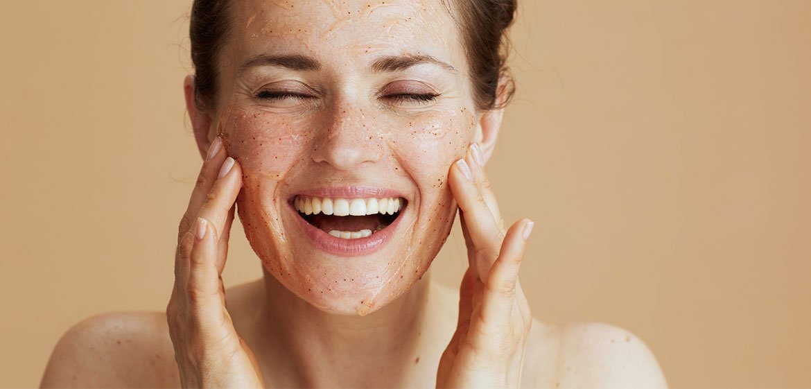 Das Gesicht einer lachenden Brünetten mit geschlossenen Augen von vorne. Sie verteilt mit beiden Händen ein leicht orangenes Peeling auf ihrem Gesicht.